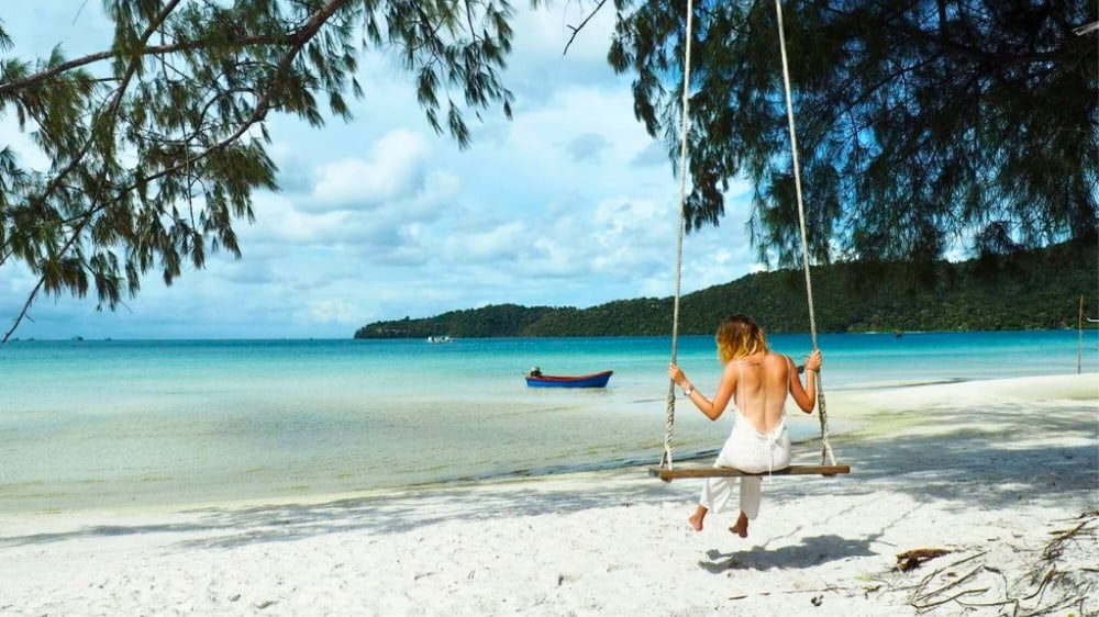 A person sitting on a rope swing hung from a pine on a Koh Rong beach, looking out over white sand and pale turquoise shallows to a small blue boat and a forested headland